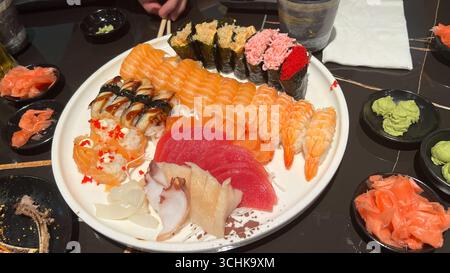 Auf einer verführerischen Sushi-Platte werden verschiedene Brötchen, frisches Sashimi und lebendige Beilagen präsentiert, die elegant auf einem runden weißen Teller präsentiert werden, um einen optischen Anblick zu erzielen Stockfoto