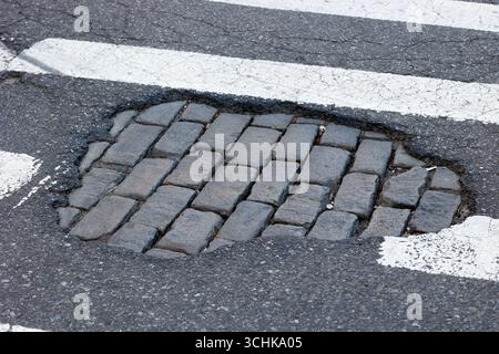 Ein New York City Crosswalk Schlagloch mit einem Stück fehlenden Asphalt offenbart eine kopfsteingepflasterte Straße darunter. Stockfoto