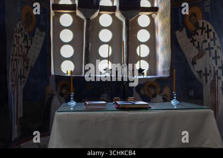 Religiöser Altar in einer alten Kirche mit Kerzenständern und Ikonen an der Wand, beleuchtet durch ein Fenster mit kleinen runden Glasscheiben Stockfoto