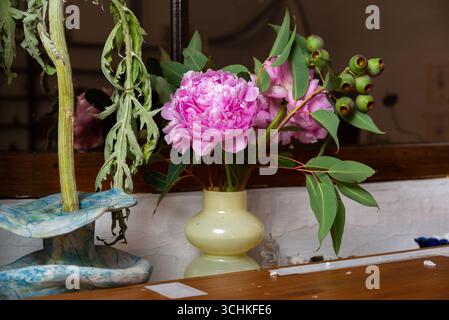 Ein bezauberndes Blumenarrangement mit leuchtenden rosafarbenen Pfingstrosen und Grün in einer einfachen Vase. Die Einrichtung bietet eine rustikale, natürliche Eleganz, die jede noch schöner macht Stockfoto