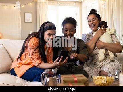 Auf der Couch teilen sich verschiedene weibliche Freunde Momente auf dem Smartphone, lächeln warm, genießen die Zeit zu Hause Stockfoto