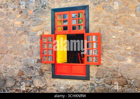 Kreativ bemaltes Fenster und Steinmauer. Leuchtendes orangefarbenes Fenster mit offenen Fensterläden, die an einer rauen Steinmauer angebracht sind. Durch das Fenster, ein heller Stockfoto