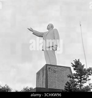 Schwarzweiß-Archivbilder aus Sloviansk, 1950er-1970er Jahre, die das Denkmal für Wladimir Lenin auf dem zentralen Revolutionsplatz (heute Soborna-Platz) darstellen. Die Statue in voller Länge mit ihrer charakteristischen Geste der zeigenden Hand ist vor einem bewölkten Himmel zu sehen. In einem Rahmen wird ein Teil des Denkmals von einem Nadelbaum verdeckt, wodurch eine komplexe Komposition entsteht. Diese Fotografien sind ein wichtiges historisches Dokument, das die sowjetische Ära im Leben der Stadt und ihre politische Symbolik vor der Entgemeinschaftung und dem Krieg im Donbass widerspiegelt Stockfoto