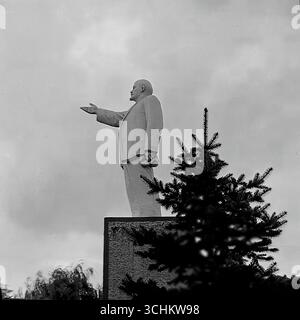 Schwarzweiß-Archivbilder aus Sloviansk, 1950er-1970er Jahre, die das Denkmal für Wladimir Lenin auf dem zentralen Revolutionsplatz (heute Soborna-Platz) darstellen. Die Statue in voller Länge mit ihrer charakteristischen Geste der zeigenden Hand ist vor einem bewölkten Himmel zu sehen. In einem Rahmen wird ein Teil des Denkmals von einem Nadelbaum verdeckt, wodurch eine komplexe Komposition entsteht. Diese Fotografien sind ein wichtiges historisches Dokument, das die sowjetische Ära im Leben der Stadt und ihre politische Symbolik vor der Entgemeinschaftung und dem Krieg im Donbass widerspiegelt Stockfoto