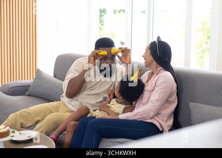 Die Familie genießt einen verspielten Moment mit Partyhüten auf dem gemütlichen Wohnzimmer-Sofa Stockfoto