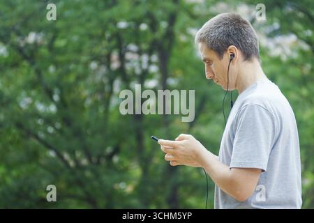 Millennial man hört pädagogische Podcasts oder Hörbuchinhalte auf einem Mobiltelefon mit Ohrhörern im grünen Park. Konzepte des lebenslangen Lernens Stockfoto