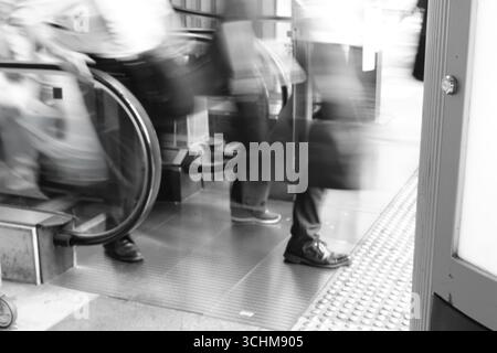 Verschwommene Geschäftsleute verlassen eine Rolltreppe in einem stark frequentierten städtischen Raum und spiegeln so den Rhythmus des beruflichen Alltags wider. Stockfoto