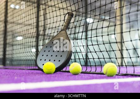 Der schwarze Padel-Schläger lehnt sich gegen das Netz auf dem violetten Rasenplatz, gelbe Bälle sind neben der Linie ausgerichtet Stockfoto