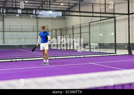Männlicher Padel-Spieler, der im Glaspadel-Platz mit Schläger neben gelbem Tennisball steht Stockfoto