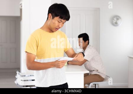 Asiatischer Teenager liest Brief, während Vater von der Küchenzeile aus zusieht Stockfoto