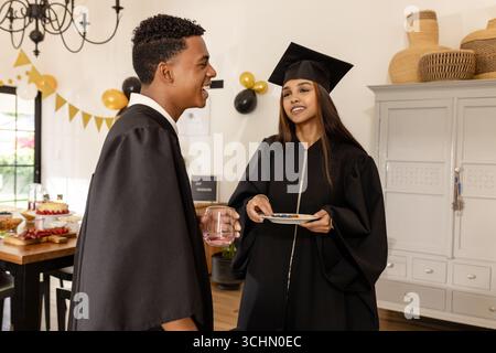 Verschiedene Absolventen in Mützen und Kleidern feiern zu Hause mit Speisen und Getränken Stockfoto