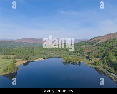Ein Blick aus der Vogelperspektive zeigt einen ruhigen See umgeben von üppigen grünen Bäumen und sanften Hügeln unter einem hellblauen Himmel. Stockfoto