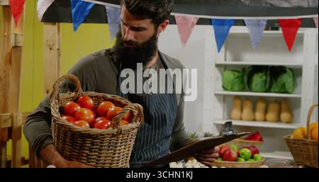 Mit Tomaten und Klemmbrett, bärtiger Verkäufer mit Schürze am Stand mit Regalfach Stockfoto