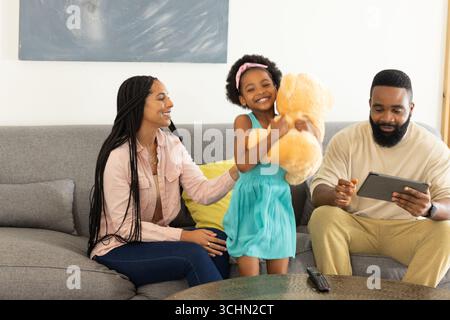 Glückliches Mädchen, das Teddybären umarmt, während Eltern sich auf der Couch mit Tablet entspannen Stockfoto