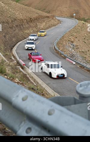 Aus nächster Nähe sehen Sie einen Sportwagenkonvoi, der durch eine kurvenreiche Bergstraßenkurve umrandet wird, die von felsigen, trockenen Hängen begrenzt wird. Stockfoto