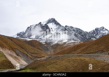 Eine gewundene Bergstraße führt über braune Hänge zu einem massiven, schneebedeckten Gipfel, der teilweise von niedrigen Wolken umhüllt ist. Stockfoto