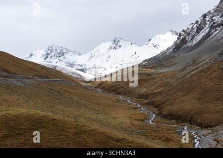 Ein sich windender Bach fließt durch ein braunes Bergtal zu hoch aufragenden, schneebedeckten Gipfeln unter bewölktem Himmel. Stockfoto