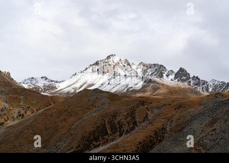 Eine malerische Bergkette mit braunen, felsigen Hängen im Vordergrund, die sich bis zu scharfen, schneebedeckten Gipfeln unter bewölktem Himmel erheben. Stockfoto