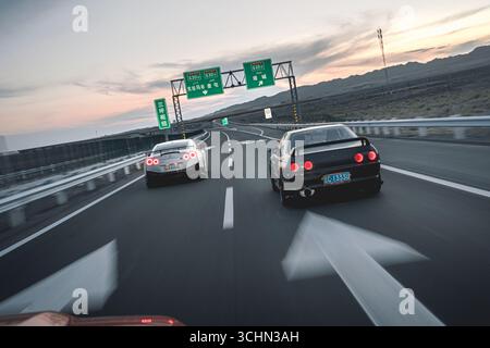 Ein weißer Nissan GT-R und eine schwarze Nissan Skyline fahren nebeneinander auf einer mehrspurigen Autobahn unter Sonnenuntergang. Stockfoto