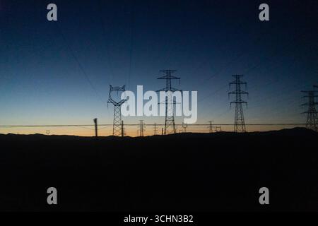 Silhouetten von Hochspannungs-Stromübertragungstürmen und -Linien vor einem farbenfrohen Sonnenuntergang über dunklen Hügeln. Stockfoto