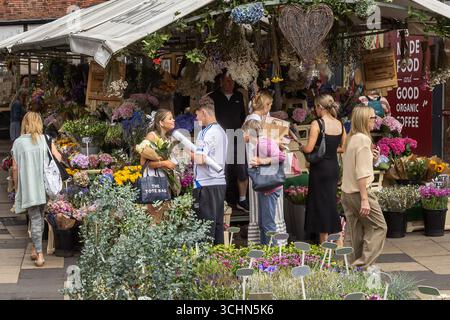 Ein belebter Blumenstand auf einem Gemeinschaftsmarkt mit lebhaften Blüten, handgefertigter Einrichtung und Shopping-Besuchern, die in einer lebhaften und einladenden Atmosphäre stöbern. Stockfoto