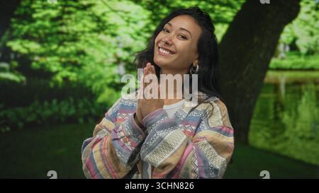 Frau mit Palmen zusammengedrückt nahe der Brust im grünen Park bei einem Baum, freundlich lächelnd und den Betrachter anschauend; Freude Dankbarkeit. Stockfoto