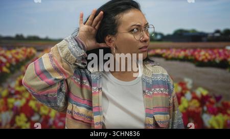 Frau, die Hand zu Ohr schröpft, hört auf einem Tulpenfeld, trägt Brille und gestreifte Jacke; Neugier-Entdeckung. Stockfoto