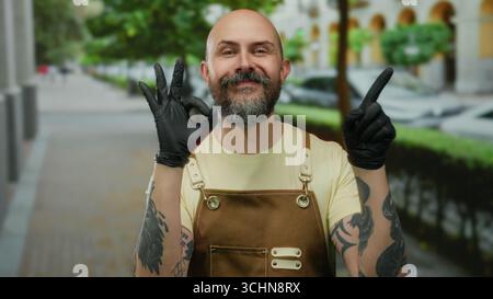 Kahler, bärtiger Mann in einer Schürze mit Tattoos und schwarzen Handschuhen, die ein OK-Zeichen auf der Stadtstraße mit verschwommenem Hintergrund zeigen. Stockfoto