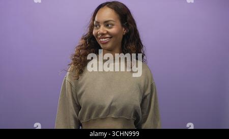 Das Gesicht der jungen Frau sieht richtig aus und trägt ein lässiges, verkürztes Sweatshirt und einen kleinen Ohrring mit Nieten im lila Studio; ruhiger Zuversicht-Optimismus. Stockfoto
