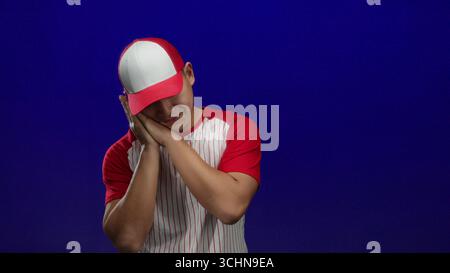 Junger chinesischer Mann in Baseballuniform mit roten und weißen Streifen, der schläfrig vor isoliertem blauem Hintergrund posiert, was auf einen müden Sportsportler hindeutet Stockfoto