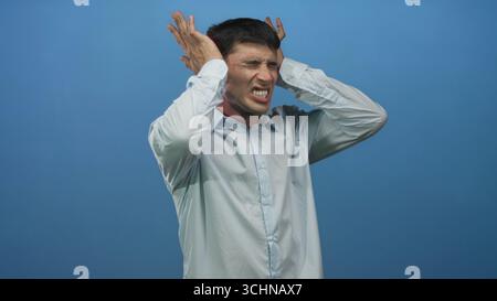 Mann, der die Hände an die Ohren drückt und ein leichtes Hemd an der blauen Studiowand trägt; Lärmbelästigung. Stockfoto
