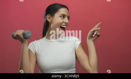 Hispanische Frau, die eine Hantel hält, während sie lächelt und vor einem leuchtend roten Hintergrund zeigt, was Fitness und Positivität zum Ausdruck bringt. Stockfoto