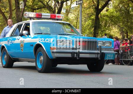 Blauer Vintage-Polizeiwagen der NYPD mit blinkenden Lichtern, der bei der Columbus Day Parade in New York City, umgeben von Menschen und Bäumen, fährt Stockfoto