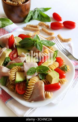 Vollkornnudelsalat mit Schinken, Avocado, Tomaten, Olivenöl und frischen Basilikumblättern auf weißem Hintergrund. Gesunde und vegetarische Lebensmittel. Sommergericht. Stockfoto