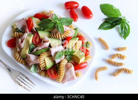Vollkornnudelsalat mit Schinken, Avocado, Tomaten, Olivenöl und frischen Basilikumblättern auf weißem Hintergrund. Gesunde und vegetarische Lebensmittel. Sommergericht. Stockfoto