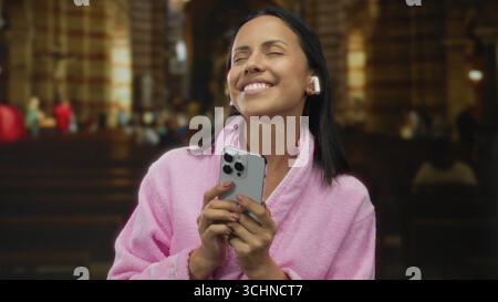 Eine Frau in rosa Bademantel mit Telefon und Ohrstöpseln steht fröhlich in einer Kirche und unterstreicht eine Mischung aus Technologie und Spiritualität. Stockfoto