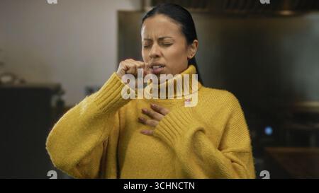 Frau hustet drinnen mit gelbem Pullover, der auf Krankheit in einem gemütlichen Raum hinweist, was einen Moment des Unbehagens und der Sorge hervorhebt. Stockfoto