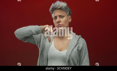 Frau mit kurzen silbernen Haaren in grauem Hoodie mit Daumen-nach-unten-Geste im roten Studio; Ablehnungsfrustration. Stockfoto