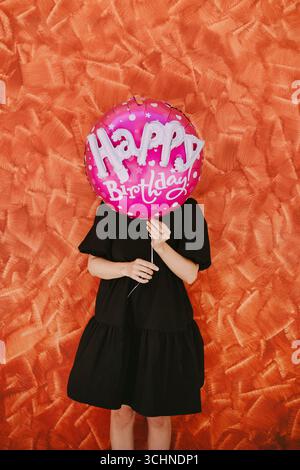 Eine Frau mittleren Alters hält einen übergroßen rosa Heliumballon mit den Worten Happy Birthday auf ihrem Gesicht, was einen verspielten und unbeschwerten Moment schafft Stockfoto