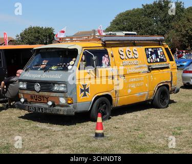 Dieses Foto zeigt einen verwitterten gelben Volkswagen T3 Van, der mit Aufklebern und einem „ROSTIGEN“ Schild verziert ist. Es hat einen Dachträger und ist auf einem Feld mit trockenem Gras geparkt. Vor dem Rad befindet sich ein kleiner Verkehrskegel, und andere Fahrzeuge und Personen sind im Hintergrund unter einem hellen, sonnigen Himmel zu sehen. Illustrativ, Editorial: Die Stokes Bay Car Rally in Gosport, Hampshire, England. 25. August 2025. Stockfoto