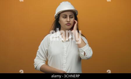 Junge hispanische Architektin mit weißem Schutzhelm und weißem Hemd, die das Ohr im orangen Studio berührt; Unwohlsein. Stockfoto