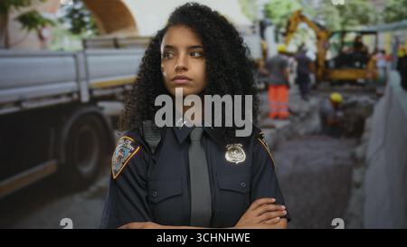 Hispanische Polizistin in Uniform steht mit gekreuzten Armen auf der Baustelle neben dem Bagger; Ablehnungssicherheit. Stockfoto