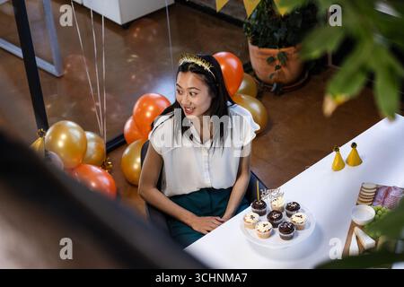 Asiatische Frau feiert Geburtstag im Büro mit Cupcakes und festlichen Ballons Stockfoto