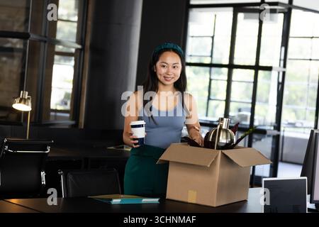 Lächelnde Frau, die Box mit Kaffeetasse in einem modernen Büroarbeitsplatz auspackt Stockfoto