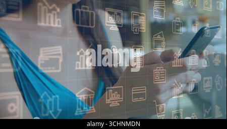 Das Smartphone in der Hand, ein Mann mit türkisfarbenem Oberteil, der Finanztechniksymbole am Schreibtisch sieht Stockfoto