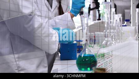 Mit Mikropipettenwissenschaftler, der Laborkittel trägt, grüne Lösung in den Kolben am Labortisch überführen Stockfoto