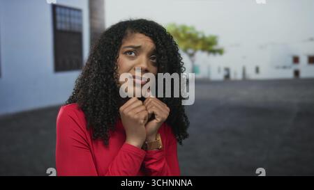 afroamerikanerin in rotem Hemd blickt durch die Finger und bedeckt das Gesicht mit den Händen auf der Straße; Angst. Stockfoto