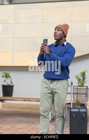 afroamerikaner, der am Bahnhof steht und Smartphone und Koffer mit Kopfhörern hält Stockfoto
