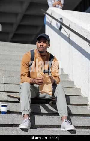 afroamerikaner sitzt auf einem Skateboard auf Betonstufen und hält braune Papiertüte, Smartphone Stockfoto
