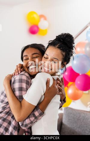 Zwei verschiedene weibliche Freunde umarmen sich zu Hause mit bunten Ballons im Hintergrund Stockfoto